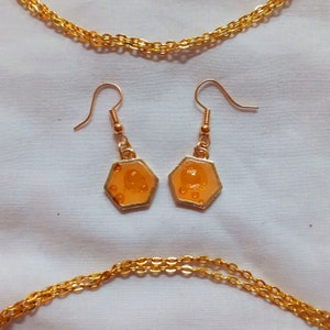 May include: A pair of gold earrings with hexagonal orange resin charms. The charms have a textured surface and a raised design. The earrings are hanging from gold hooks.