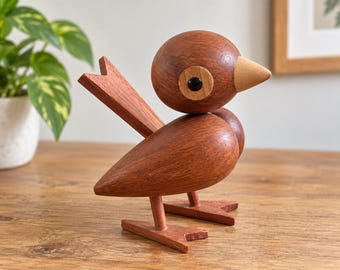 以下が含まれることがあります： 赤茶色の木材で作られた小さな鳥の置物。鳥は丸みを帯びた体、明るい色の尖ったくちばし、中央が黒い円形の目のディテールが特徴です。2本の平らで角張った足で立ち、三角形の尾を持っています。