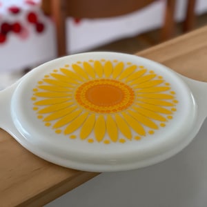 Op de afbeelding: Witte glazen deksel met een geel en oranje zonnebloemontwerp. De deksel heeft aan elke kant een handvat.