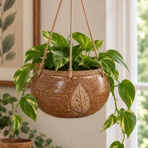 May include: A round, speckled brown ceramic hanging planter with a textured leaf imprint on the front. The planter is suspended by three natural jute ropes and contains a trailing pothos plant with variegated green and cream-colored leaves.