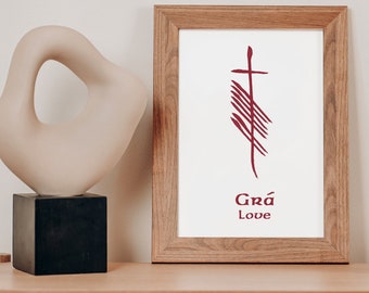 May include: A framed print with a red stylized cross and the text "Grá Love" in a wooden frame on a light wood surface.