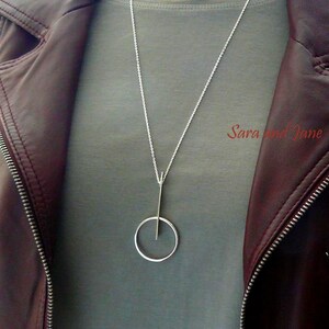 May include: A silver necklace with a long, thin pendant that has a circle at the bottom. The necklace is being worn by a person wearing a gray shirt and a brown leather jacket.