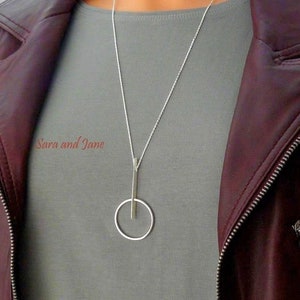 May include: A silver necklace with a long, thin pendant that forms a circle with a vertical line through it. The necklace is worn by a person wearing a gray shirt and a maroon leather jacket.