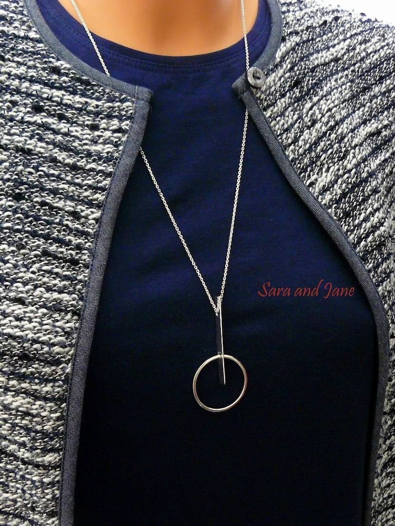 May include: A silver necklace with a circle pendant and a vertical bar intersecting the circle. The necklace is worn by a person wearing a blue shirt and a gray cardigan. The text "Sara and Jane" is visible in the image.