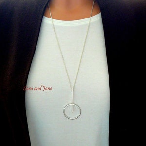 May include: A silver necklace with a long, thin pendant that forms a circle with a vertical line through it.