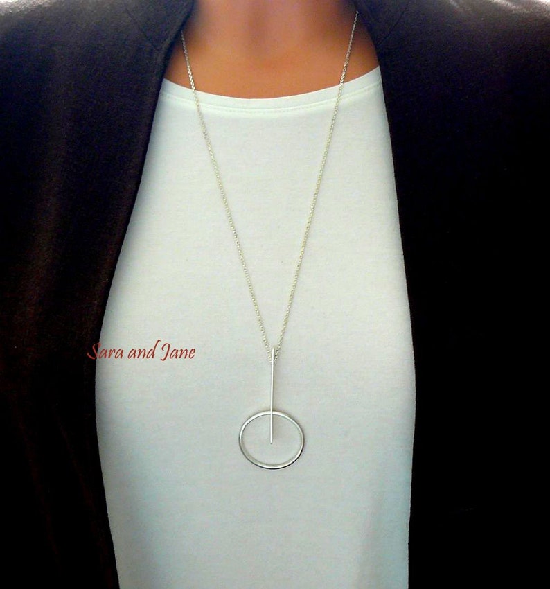 May include: A silver necklace with a long, thin pendant that forms a circle with a vertical line through it.