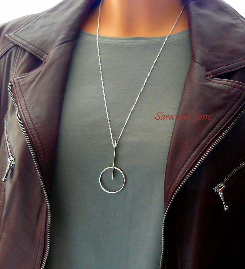 May include: A silver necklace with a long, thin pendant that has a circle at the bottom. The necklace is being worn by a person wearing a gray shirt and a brown leather jacket.