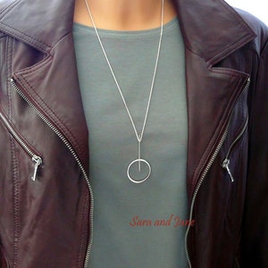 May include: A silver necklace with a thin chain and a simple circle pendant. The necklace is worn by a person wearing a gray shirt and a brown leather jacket.