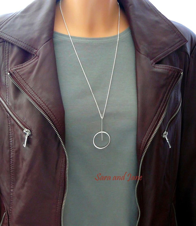 May include: A silver necklace with a thin chain and a simple circle pendant. The necklace is worn by a person wearing a gray shirt and a brown leather jacket.