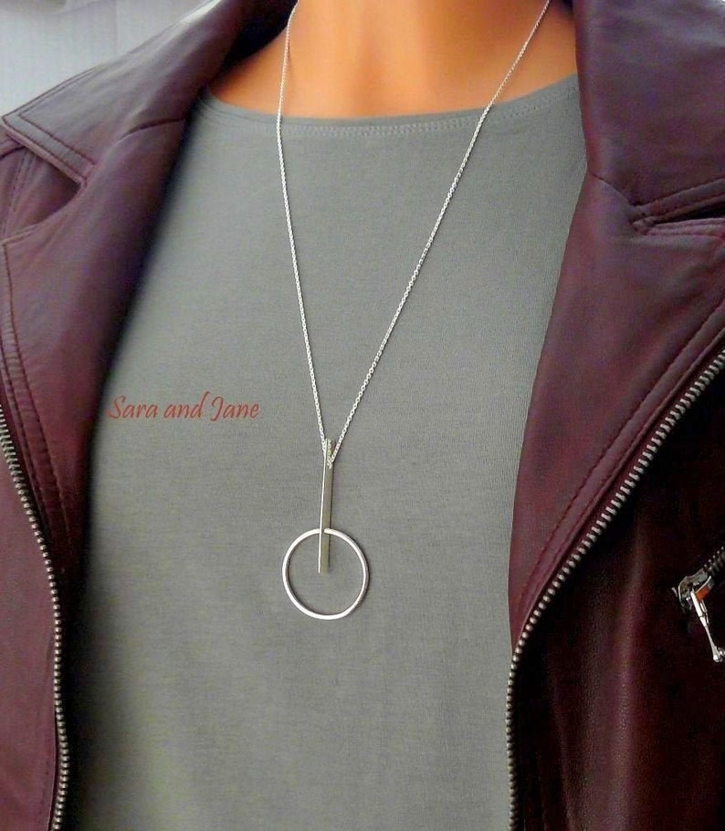 May include: A silver necklace with a long, thin pendant that forms a circle with a vertical line through it. The necklace is worn by a person wearing a gray shirt and a maroon leather jacket.