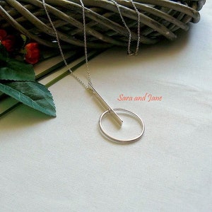 May include: A silver necklace with a long, thin bar pendant that intersects a circle pendant. The necklace is on a white fabric background.