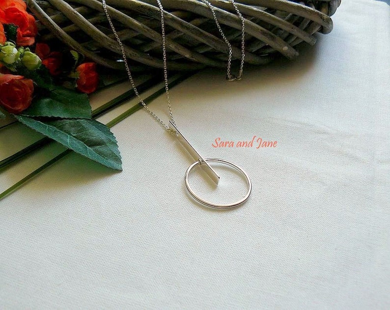 May include: A silver necklace with a long, thin bar pendant that intersects a circle pendant. The necklace is on a white fabric background.