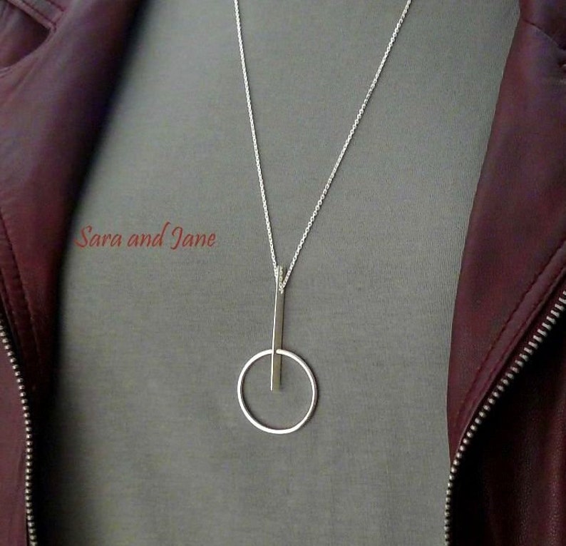 May include: A silver necklace with a long, thin pendant that forms a circle with a vertical line through it.
