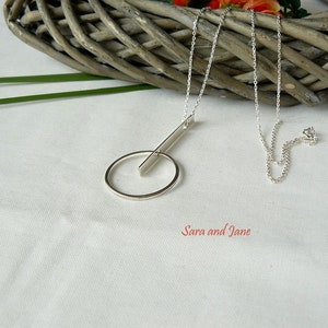 May include: A silver necklace with a circle and a bar pendant. The necklace is on a white surface with a wicker basket in the background.