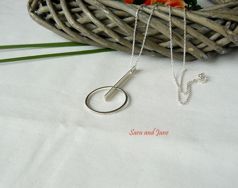 May include: A silver necklace with a circle and a bar pendant. The necklace is on a white surface with a wicker basket in the background.