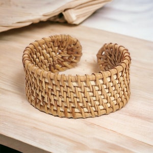 May include: A woven rattan bangle bracelet with a natural brown color. The bracelet is made of thin strips of rattan that are woven together in a tight pattern.