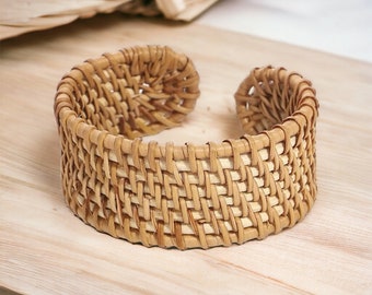 May include: A woven rattan bangle bracelet with a natural brown color. The bracelet is made of thin strips of rattan that are woven together in a tight pattern.