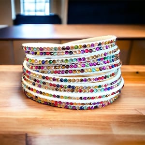 May include: A white leather bracelet with multiple rows of colorful rhinestones. The bracelet is stacked and shows the different colors of the rhinestones.