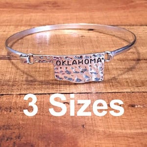May include: Silver bracelet with a hammered metal charm shaped like the state of Oklahoma. The charm has the word "OKLAHOMA" engraved on it. The bracelet is available in three sizes.