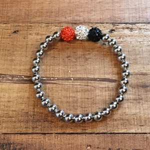 May include: A silver beaded bracelet with three accent beads: orange, white, and black.