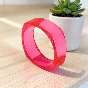 May include: A bright, translucent hot pink bangle bracelet with a smooth, rounded square shape. The bracelet is constructed from a clear, vibrant resin material that allows light to pass through, creating a vivid and saturated color effect.
