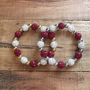 May include: Two beaded bracelets with red and white sparkly beads. The bracelets are intertwined and have a gold accent.