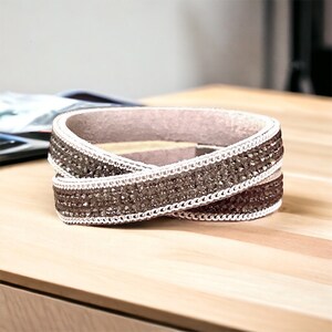 May include: A brown leather bracelet with a white and silver rhinestone trim. The bracelet is wrapped in a criss-cross pattern.