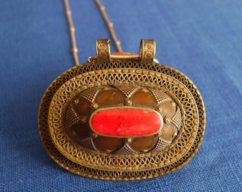 Puede incluir: Un collar con colgante antiguo de color plateado con forma ovalada. El colgante presenta una piedra roja en el centro, rodeada de intrincados filigranas y trabajos decorativos en metal. El collar cuelga de una delicada cadena sobre un fondo azul.