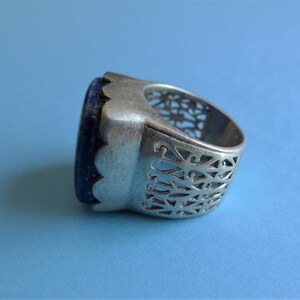 May include: A silver ring with a blue gemstone set in a scalloped bezel. The band is intricately carved with a repeating pattern.
