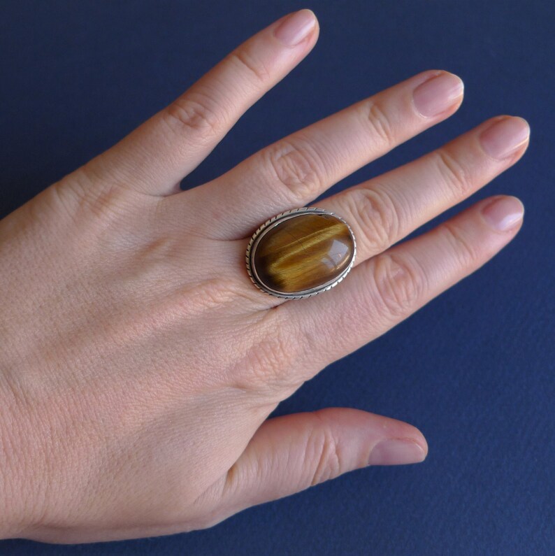 K&ouml;nnte beinhalten: Ein silberner Ring mit einem gro&szlig;en ovalen Tigerauge-Edelstein. Der Ring hat ein dekoratives silbernes Band.