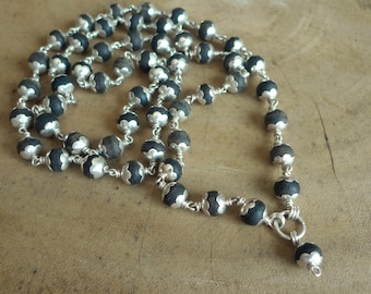 May include: A necklace featuring dark gray beads with silver-colored accents. The beads are strung together with silver-colored wire, creating a delicate and intricate design. The necklace is displayed on a wooden surface.