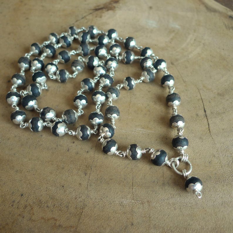 May include: A necklace with dark grey beads and silver-coloured detailing. The beads are strung together with silver wire, forming an intricate design. The necklace is displayed on a wooden surface.