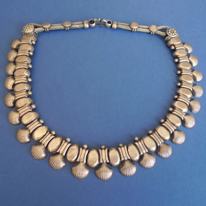 May include: Silver necklace with an intricate design. The necklace features oval and shell-shaped elements linked by a chain. The piece is displayed against a blue background, highlighting its metallic sheen and detailed craftsmanship.