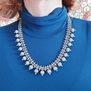 May include: A silver necklace with a chain design and round, textured beads. The necklace is worn by a person with a blue turtleneck shirt.