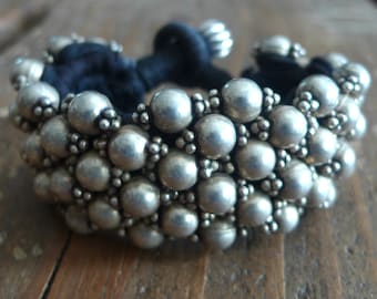 May include: A silver bracelet with a black cord and multiple rows of round beads. The beads are silver and have a textured surface.