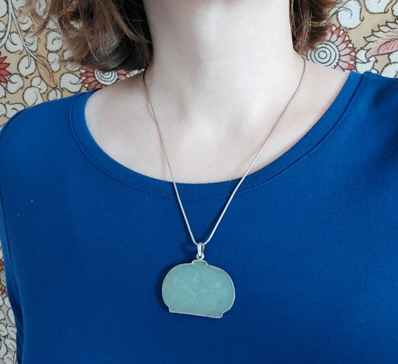 May include: A silver chain necklace with a light green pendant shaped like a vase with a floral design.