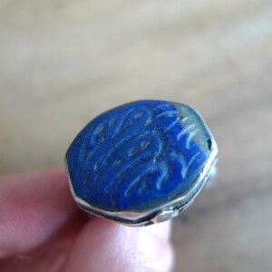 Pu&ograve; includere: Un anello d'argento con una pietra di lapislazzuli blu incastonata in una forma esagonale. La pietra presenta intricate incisioni sulla sua superficie.
