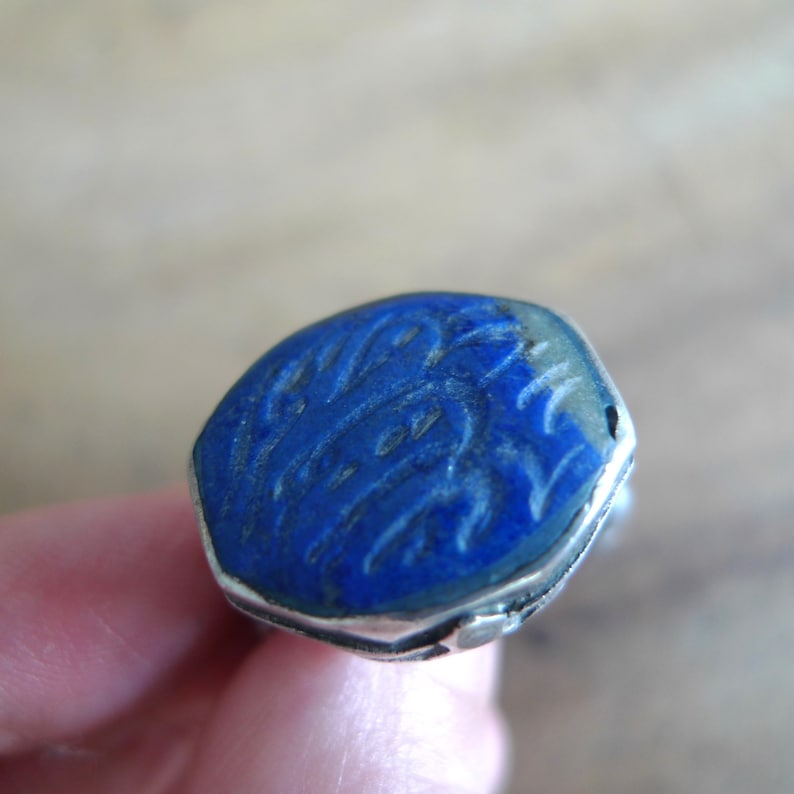 Pu&ograve; includere: Un anello d'argento con una pietra di lapislazzuli blu incastonata in una forma esagonale. La pietra presenta intricate incisioni sulla sua superficie.