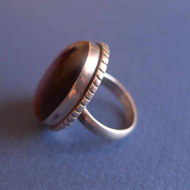 K&ouml;nnte beinhalten: Ein silberner Ring mit einem braunen ovalen Edelstein, der in einer strukturierten Fassung sitzt.