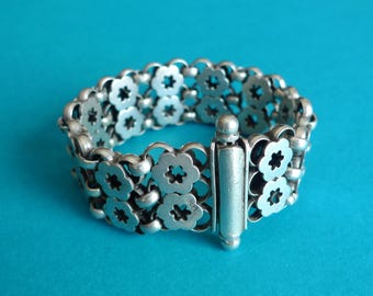 May include: A silver-toned bracelet with a floral design. The bracelet features multiple rows of interconnected flower-shaped links, creating a flexible band. The clasp is a rectangular bar with a small, rounded end. The background is a solid teal color.