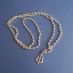 Puede incluir: Un collar de tono plateado que presenta una cadena larga de pequeñas cuentas esféricas uniformes. El collar está adornado con un colgante único con forma de tridente de tres puntas, que cuelga de la parte inferior de la cadena de cuentas.