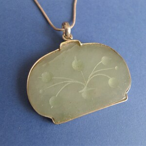 May include: A silver chain necklace with a green jade pendant. The pendant is oval shaped and has a carved floral design.
