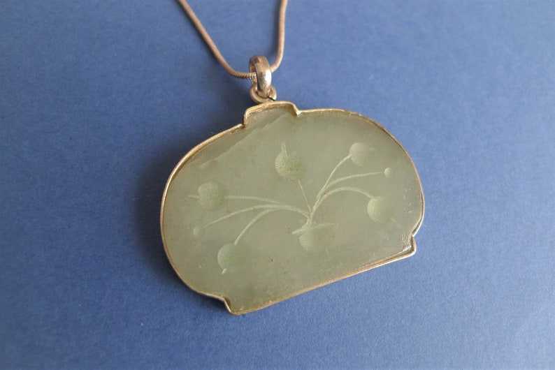 May include: A silver chain necklace with a green jade pendant. The pendant is oval shaped and has a carved floral design.