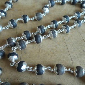 May include: Close-up of a beaded necklace featuring dark, round beads and silver-coloured accents. The beads are strung together with a delicate silver chain, creating a detailed and elegant design. The necklace is displayed on a wooden surface.