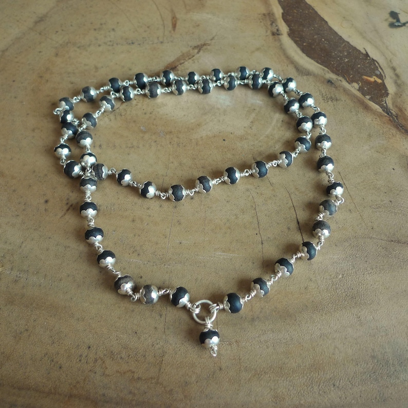 May include: A layered necklace with dark grey beads and silver metal detailing. The necklace has a delicate chain design with a small pendant at the bottom. The beads are evenly spaced, creating a symmetrical pattern.