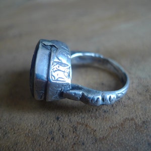 Pu&ograve; includere: Un anello d'argento con una pietra preziosa blu incastonata in una lunetta quadrata. L'anello &egrave; decorato con intricati motivi floreali e ha una fascia larga.