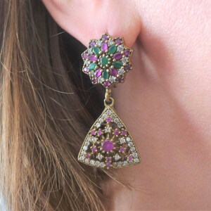 May include: A pair of dangle earrings with a star-shaped top and a triangle-shaped bottom. The earrings are made of gold-toned metal and are adorned with clear, pink, and green gemstones.