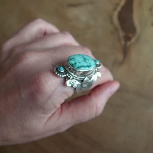 May include: A silver ring with a large, oval turquoise stone. The ring has detailed silverwork around the main stone and smaller turquoise accents. The ring is on a hand.