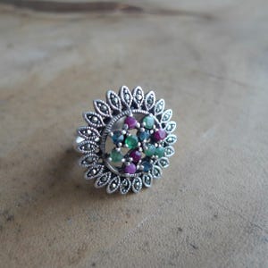 Può includere: Un anello d'argento con un design floreale. L'anello presenta un gruppo centrale di pietre preziose colorate, tra cui pietre rosse, verdi e blu, circondato da un bordo d'argento decorativo. L'anello è posto su uno sfondo neutro.