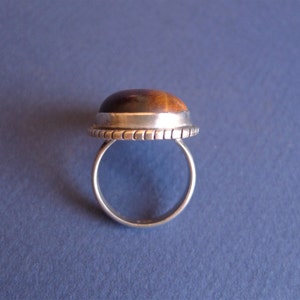 K&ouml;nnte beinhalten: Ein silberner Ring mit einem braunen und goldenen Tigerauge-Stein, der in einer Zargenfassung gefasst ist. Der Ring hat eine strukturierte Band.